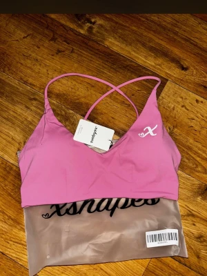 Rosa sport-bh med tunna axelband - Snygg rosa sport-bh från xshapes med tunna korsade axelband i ryggen och liten vit logga framtill. Tillverkad i ett stretchigt och mjukt material som passar perfekt för träning eller yoga.