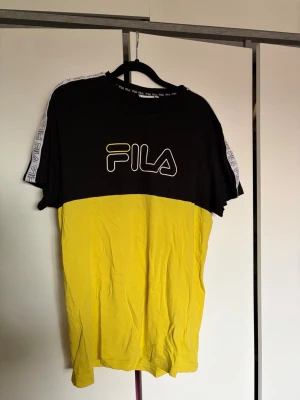 Svart och gul FILA t-shirt  - Säljer en snygg svart och gul t-shirt från FILA i storlek XL. T-shirten har korta ärmar med vita FILA-loggor längs axlarna och ett stort FILA-tryck på bröstet. 