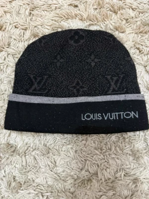 Svart Louis Vuitton mössa med logga - Snygg svart mössa från Louis Vuitton med klassiskt monogrammönster och grå kant. Broderad logga framtill. Perfekt accessoar för att lyfta din stil under kyliga dagar.
