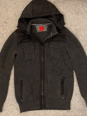Mörkgrå stickad jacka med huva - Säljer en mörkgrå stickad cardigan med dragkedja och huva. Jackan har ribbad struktur, två bröstfickor med knappar och detaljer i syntetmaterial över axlar och huva. Perfekt för vår/höst dagar och har en casual look. Skriv för frågor! 