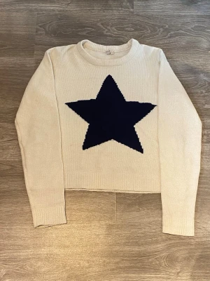 Beige stickad tröja med mörkblå stjärna - Snygg beige stickad tröja med en stor mörkblå stjärna på bröstet. Tröjan har rund halsringning, långa ärmar och ribbade muddar. Materialet är mjukt och skönt.