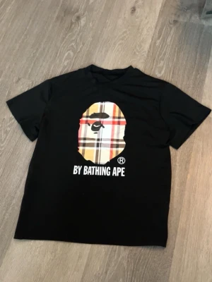Burberry collab med bathing by ape tröja - Svart t-shirt med klassiskt BAPE-tryck framtill i rutigt mönster och texten 'BY BATHING APE' under. T-shirten har rund hals och korta ärmar, perfekt för en streetwear-look.
