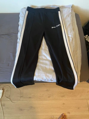 Palm Angels svarta mjukisbyxor - Palm Angels svarta mjukisbyxor med vita sidoränder och dragkedja vid benslut. Byxorna har resår i midjan och logga tryckt på vänster sida. Sköna och stilrena, perfekta för en avslappnad look.