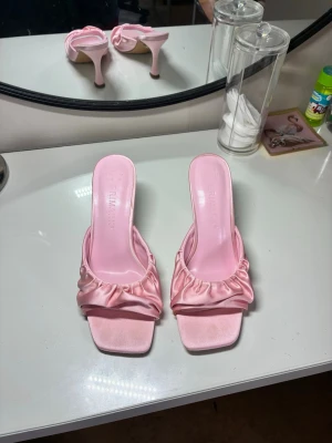 Rosa satin-klackskor med rynkad rem - Säljer ett par ljusrosa pumps i satin med öppen tå och fyrkantig framdel. Skorna har en rynkad rem över foten och smal klack. Perfekta för dig som vill ha en söt och trendig look med extra detaljer. Klacken är medelhög och designen känns både modern och feminin.