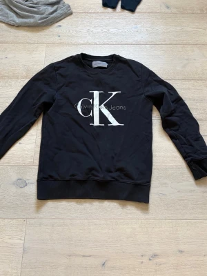 Svart sweatshirt från Calvin Klein - Svart sweatshirt från Calvin Klein Jeans med stort vitt CK-tryck på bröstet. Tröjan har rund halsringning, långa ärmar och ribbade muddar vid ärmslut och nederkant. Perfekt för en avslappnad och stilren look.
