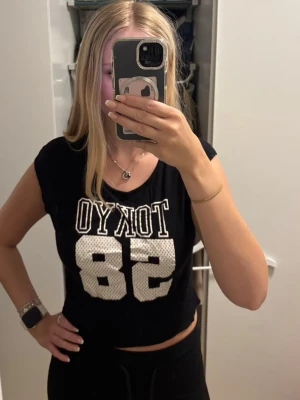 Svart croppad topp TOKYO 58 Divided - Säljer en svart croppad topp från Divided med vit text 'TOKYO 58' framtill. Toppen har korta ärmar och en loose passform, perfekt för en avslappnad streetstyle-look. Materialet är mjukt och skönt, troligen bomull eller bomullsblandning.