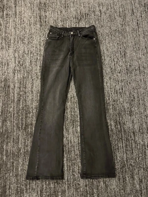 Svarta bootcut jeans med fyra fickor - Säljer ett par svarta bootcut jeans med klassisk femficksdesign och dragkedja med knapp. Jeansen har lätt utsvängda ben och subtila slitningar framtill. Materialet är denim med en mjuk känsla och sömmar i matchande färg.