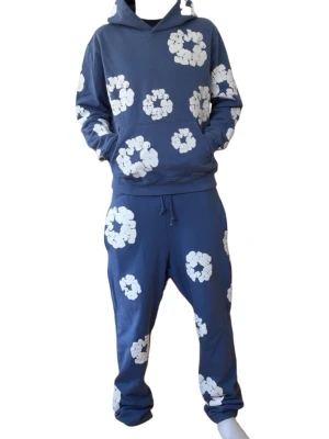 Blå denim tears tracksuit med vita blommor - Säljer en blå hoodie med huva och stora vita blommönster över hela tröjan. Hoodien har en avslappnad passform och är gjord i mjukt bomullstyg. Perfekt för dig som gillar färgglada och unika plagg med skön streetwear-vibe.
