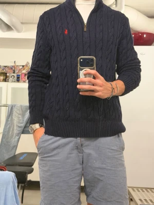 Mörkblå kabelstickad tröja Polo Ralph Lauren - Mörkblå kabelstickad tröja från Polo Ralph Lauren med halv dragkedja och röd broderad logga på bröstet. Tröjan har ribbade muddar och hög krage, perfekt för lager på lager. Klassisk och tidlös design i mjukt material. Passar mera som L