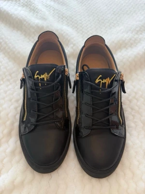 Svarta sneakers med gulddetaljer - Säljer ett par äkta svarta Giuseppe Zanotti med lackdetaljer och guldfärgade dragkedjor på sidorna. Skorna har snörning och en cool guldfärgad logga på plösen. Perfekta för dig som gillar en stilren men ändå lyxig look. Kvitto finns ej kvar men när man får skorna kan man alltid lämna tillbaka om man ångrar sig