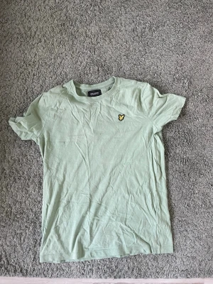 Ljusgrön t-shirt från Lyle & Scott - Snygg ljusgrön t-shirt från Lyle & Scott med klassisk rund hals och korta ärmar. På bröstet sitter den ikoniska gula loggan som detalj. Tillverkad i mjuk bomull för skön känsla hela dagen.