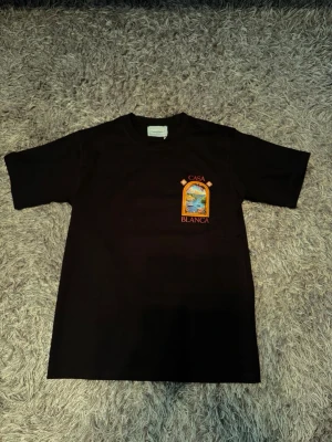 Svart Casa Blanca t-shirt med tryck - Snygg svart t-shirt från Casa Blanca med färgglatt tryck av en hamn och blommor både fram och bak. T-shirten har korta ärmar och rund hals. Perfekt för dig som vill sticka ut med ett unikt motiv och cool vibe.