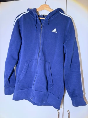 Blå zip hoodie från Adidas - Blå hoodie från Adidas med dragkedja, vit logga på bröstet och klassiska vita ränder längs ärmarna. Utrustad med huva och känguruficka framtill. Tillverkad i mjukt sweatshirtmaterial, perfekt för en sportig och avslappnad stil.