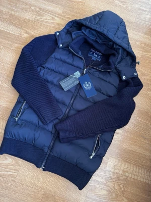 Cain - Snygg mörkblå dunväst från Mason's med avtagbar huva och stickade ärmar. Västen har två dragkedjeförsedda fickor och ribbade muddar. Materialet är en mix av dun och stickat tyg, vilket ger en modern och sportig look.