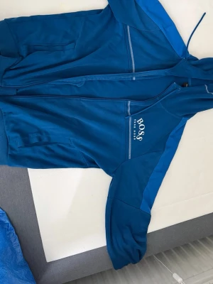 Blå hoodie från BOSS med dragkedja - Snygg blå hoodie från BOSS med dragkedja framtill och justerbar huva. Hoodien har ribbade muddar, fickor på sidorna och BOSS-logga på bröstet. Tillverkad i mjukt material som känns skönt mot huden. Perfekt för en avslappnad och sportig stil.