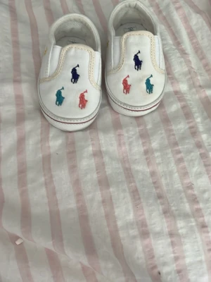 Vita babyskor med färgglada loggor - Säljer ett par vita babyskor från Ralph Lauren med broderade loggor i blått, rött, lila och turkost framtill. Skorna har elastiska sidor för enkel påtagning och mjuk sula med rosa Ralph Lauren-tryck under. Perfekta för små trendsetters!
