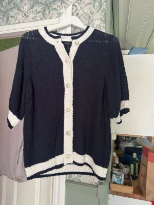Marinblå kofta med vita detaljer - Snygg marinblå kofta med vita kontrastkanter och pärlemorknappar. Koftan har halvlånga ärmar och ribbade muddar. Perfekt till jeans eller kjol för en klassisk look. Materialet är stickat och känns lätt och luftigt.