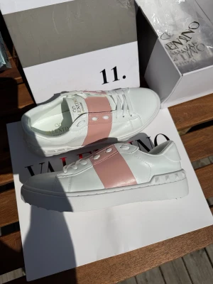 Valentino Garavani vita sneakers - Snygga vita sneakers från Valentino Garavani med bred ljusrosa rem över vristen och nitar på sidan. Skorna är i läder med klassisk låg siluett och tjock vit sula. Perfekta för dig som vill ha en clean men ändå unik look. Båda skorna är vänster, därav priset 