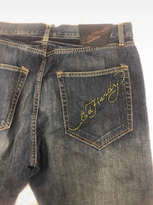 Mörkblå Ed Hardy jeans med brodyr - Säljer ett par mörkblå jeans från Ed Hardy med gul broderad logga på bakfickan. Jeansen har raka ben, klassisk femficksmodell och slitningar för en cool vintage-look. Perfekta för dig som gillar streetwear och vill sticka ut.