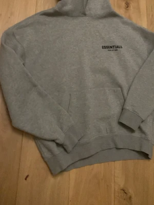 Grå Essentials Fear of God hoodie - Säljer en grå hoodie från Essentials Fear of God med svart logga på bröstet. Klassisk passform med huva och magficka. Tillverkad i mjukt material som känns skönt mot huden. Perfekt för dig som gillar streetwear och enkel stil.