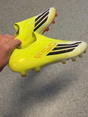 Gula fotbollsskor med svarta ränder - Säljer ett par riktigt snygga fotbollsskor i neon-gult med svarta och vita detaljer samt röda inslag på sulan. Skorna har låg profil och är designade för fart på planen. Ovandelen är i syntetmaterial och sulan har fasta dobbar för spel på gräs.