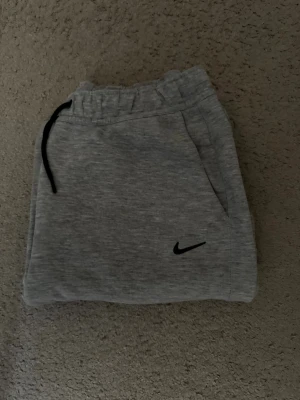 Gråa mjukisbyxor från Nike - Snygga grå mjukisbyxor från Nike med svart dragsko i midjan och diskret Nike-logga på benet. Byxorna har raka ben, sidofickor och är tillverkade i mjukt sweatshirtmaterial. Perfekta för chill eller träning.