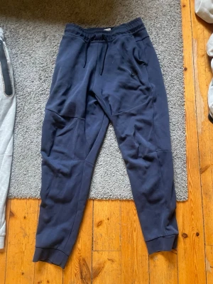Mörkblå mjukisbyxor från Nike - Säljer ett par mörkblå mjukisbyxor från Nike med dragsko i midjan och ribbade muddar vid bensluten. Byxorna har sidofickor och en broderad Nike-logga på benet. Perfekta för chill dagar eller träning. ( funkar för storlek S-M)