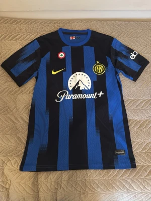 Inter Milan tröja - Säljer en officiell Inter Milan matchtröja med blå och svarta ränder, Nike-logga och Paramount+ sponsortryck. Tröjan har korta ärmar och klubbmärke på bröstet. Perfekt för dig som älskar fotboll och vill bära ditt lag med stil.