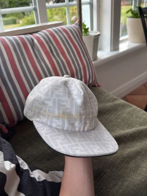Vit keps med Fendi-logga - Snygg vit keps från Fendi med det klassiska FF-mönstret över hela kepsen. Justerbar baktill för perfekt passform och tillverkad i ett mjukt tygmaterial. Perfekt för dig som vill ha en stilren accessoar med lyxig känsla.