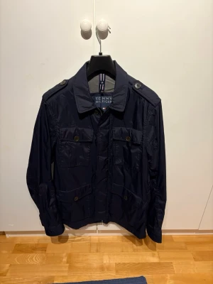 Marinblå jacka från Tommy Hilfiger - Snygg marinblå jacka från Tommy Hilfiger med klassisk krage, fyra framfickor med knappar och axelklaffar. Jackan har dragkedja och knappar framtill och är tillverkad i lätt polyester, perfekt för vår och höst.