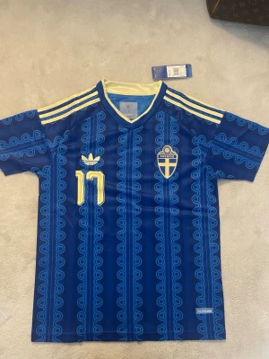 Blå Sverige fotbollströja Gyökeres  - Säljer en blå fotbollströja från Sverige med gula detaljer och nummer 17. Tröjan har korta ärmar, V-ringning och mönstrade ränder. På bröstet finns Sveriges landslagsemblem och Adidas-logga. Namnet Gyökeres står på ryggen.