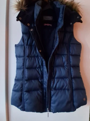 Mörkblå dunväst med fuskpäls Lee Cooper  - Snygg mörkblå dunväst med avtagbar huva och fuskpälsdetalj. Västen har quiltad design, dragkedja och tryckknappar framtill samt två sidofickor. Perfekt för lager-på-lager och ger en sportig look.