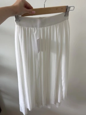 Vit långkjol - Säljer en vit långkjol med bred midjeresår och lätt plisserad look. Oanvänd och prislapp kvar. Ordinarie pris 349kr.