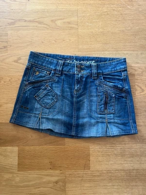 Blå jeanskjol med fickor och bälte - Säljer en blå jeanskjol i kort modell med flera coola fickor, dragkedjedetalj och dekorativa nitar. Kjolen har ett matchande jeansbälte och slitna detaljer för en avslappnad look. Perfekt för dig som gillar streetstyle och denim.