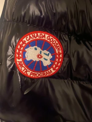Svart dunväst från Canada Goose - DRAGKEDJAN ÄR SÖNDRIG!!! Snygg svart dunväst från Canada Goose med klassisk röd och vit logotyp på bröstet. Västen har glansigt material, quiltad design och dragkedja framtill. Perfekt för lager-på-lager och håller dig varm under kyliga dagar.