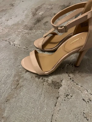 Beige högklackade sandaler - Snygga beige sandaler med smal rem över tårna och justerbar ankelrem med spänne. Skorna har hög klack och öppen tå, vilket ger en elegant och stilren look. Tillverkade i ett glansigt material som ger en fräsch finish.