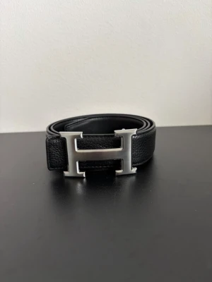 Svart skärp med stor H-spänne - Snyggt svart bälte i skinn med ett stort, silverfärgat H-format spänne. Perfekt accessoar för att lyfta vilken outfit som helst. Klassisk design som funkar till många olika stilar.