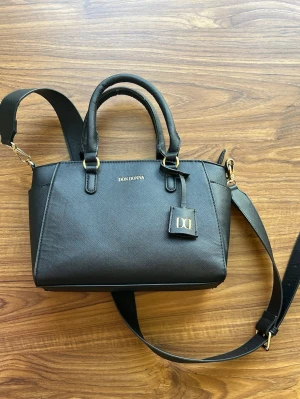 Svart handväska från Don Donna - Snygg svart handväska från Don Donna i stilren design. Väskan har två korta handtag och en avtagbar axelrem i matchande färg. Guldiga metalldetaljer och logga framtill. Tillverkad i ett tåligt konstläder med struktur. Perfekt för dig som gillar klassisk och trendig stil.