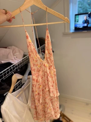 Blommig kort sommarklänning - Söt kort klänning med blommigt mönster i rosa, rött och gult. Klänningen har tunna axelband, v-ringning och volangdetaljer framtill. Perfekt för varma dagar och har en luftig, lätt känsla.