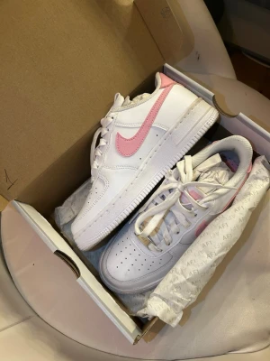 Nike Air Force 1 vit/rosa sneakers - Säljer ett par Nike Air Force 1 sneakers i vitt med rosa swoosh. Klassisk låg modell med perforerad tå, vita skosnören och robust sula. Perfekta för dig som gillar stilrena och fräscha sneakers med en touch av färg.(  Använd 1 gång )