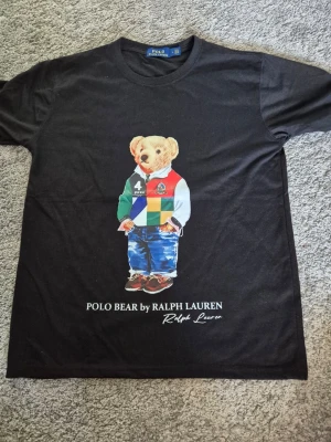 Svart Polo Bear t-shirt Ralph Lauren - Svart t-shirt från Ralph Lauren med ikoniska Polo Bear-trycket framtill. Björnen har färgglada kläder och trycket är detaljerat. Klassisk rund hals och normal passform. Perfekt för dig som gillar streetwear och vill sticka ut lite extra.