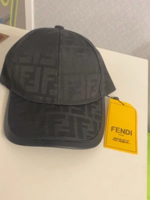 Svart keps med Fendi-logga - Snygg svart keps från Fendi med det klassiska FF-mönstret över hela kepsen. Kepsen har böjd skärm och justerbar baksida. Tillverkad i ett slitstarkt tyg med Fendi-detaljer även på insidan. Perfekt för dig som gillar exklusiva accessoarer.