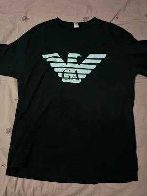 Svart t-shirt med stor logga - Storlek L Snygg svart t-shirt med stor vit logga framtill i form av en örn och bokstäverna GA. Klassisk passform och rund halsringning. Perfekt till jeans eller shorts för en clean och stilren look.