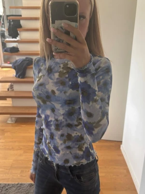 Blommig meshtröja från Lindex - Säljer en tunn, långärmad meshtröja från Lindex med blått och grönt blommönster på ljus bakgrund. Tröjan har figurnära passform och vågig kant vid ärmslut och nederkant. Perfekt för att addera färg till din outfit!