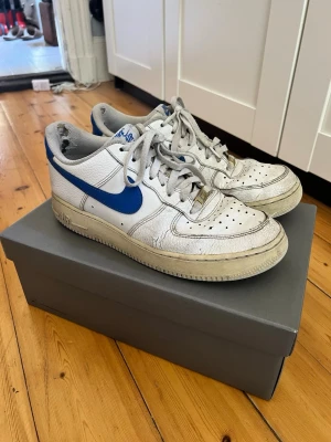 Nike Air Force 1 vita/blå sneakers - Klassiska Nike Air Force 1 sneakers i vitt läder med blå swoosh och detaljer. Lågt skaft, perforerad tåbox och vita skosnören. Guldiga AF1-detaljer på snörningen och grå yttersula. Perfekta för dig som gillar retrostil och streetwear.