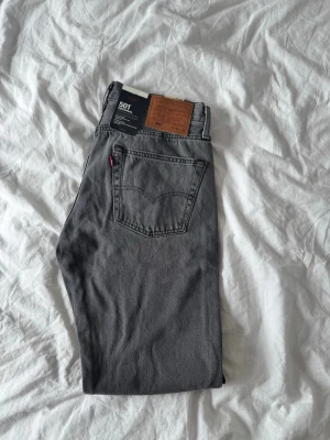 Grå Levis 501 straight jeans - Säljer ett par klassiska grå Levis 501 jeans med raka ben och normal passform. Jeansen har fem fickor, knappgylf och den ikoniska Levis-lappen bak. Perfekta för dig som gillar en tidlös look och vill ha ett par jeans som funkar till allt.