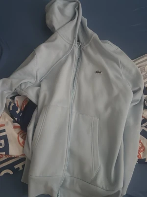 Ljusblå hoodie med dragkedja - Snygg ljusblå hoodie från Lacoste med dragkedja framtill och klassisk krokodillogga på bröstet. Hoodien har huva med dragsko och två stora fickor framtill. Perfekt för en avslappnad och trendig stil.