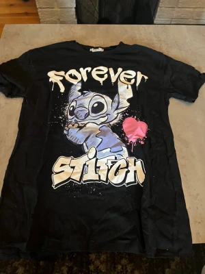Svart Stitch t-shirt från Disney - Cool svart t-shirt från Disney med stort tryck av Stitch och texten 'Forever Stitch' i graffiti-stil. T-shirten har en rosa hjärtdetalj och är kortärmad. Perfekt för dig som älskar Lilo & Stitch och vill ha en unik och lekfull look.
