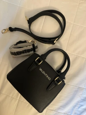 Svart handväska från Valentino - Valentino väska! En lyxig och verkligen vacker väska men jag känner att den guldfärgade detaljen inte passar mig, så därför säljer jag den. Nypriset var 1346 kr. Det kan finnas mycket små spår av användning men väskan är fortfarande i fint skick. Jag packar den noggrant innan leverans!🌷