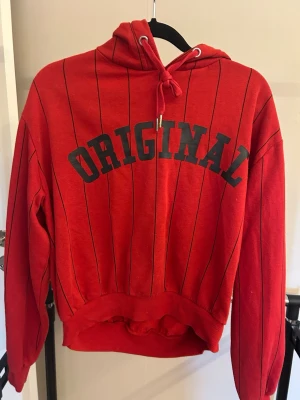 Röd hoodie med svart text - Röd hoodie med svarta tunna ränder och texten 'ORIGINAL' i svart över bröstet. Tröjan har huva med dragsko och ribbade muddar vid ärmar och midja. 